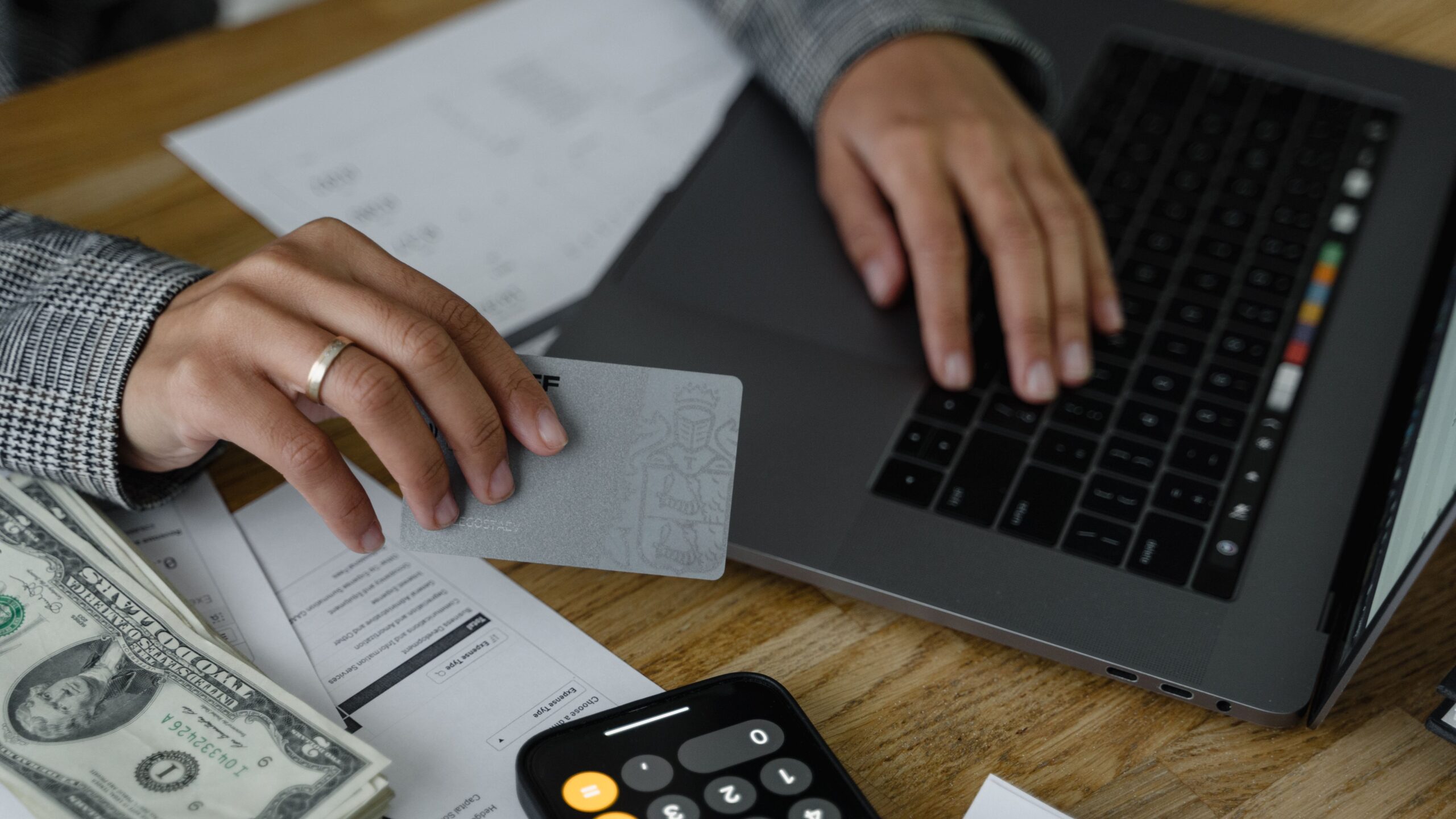 Close-up of a person using a credit card and laptop in a financial setting with cash and calculator.