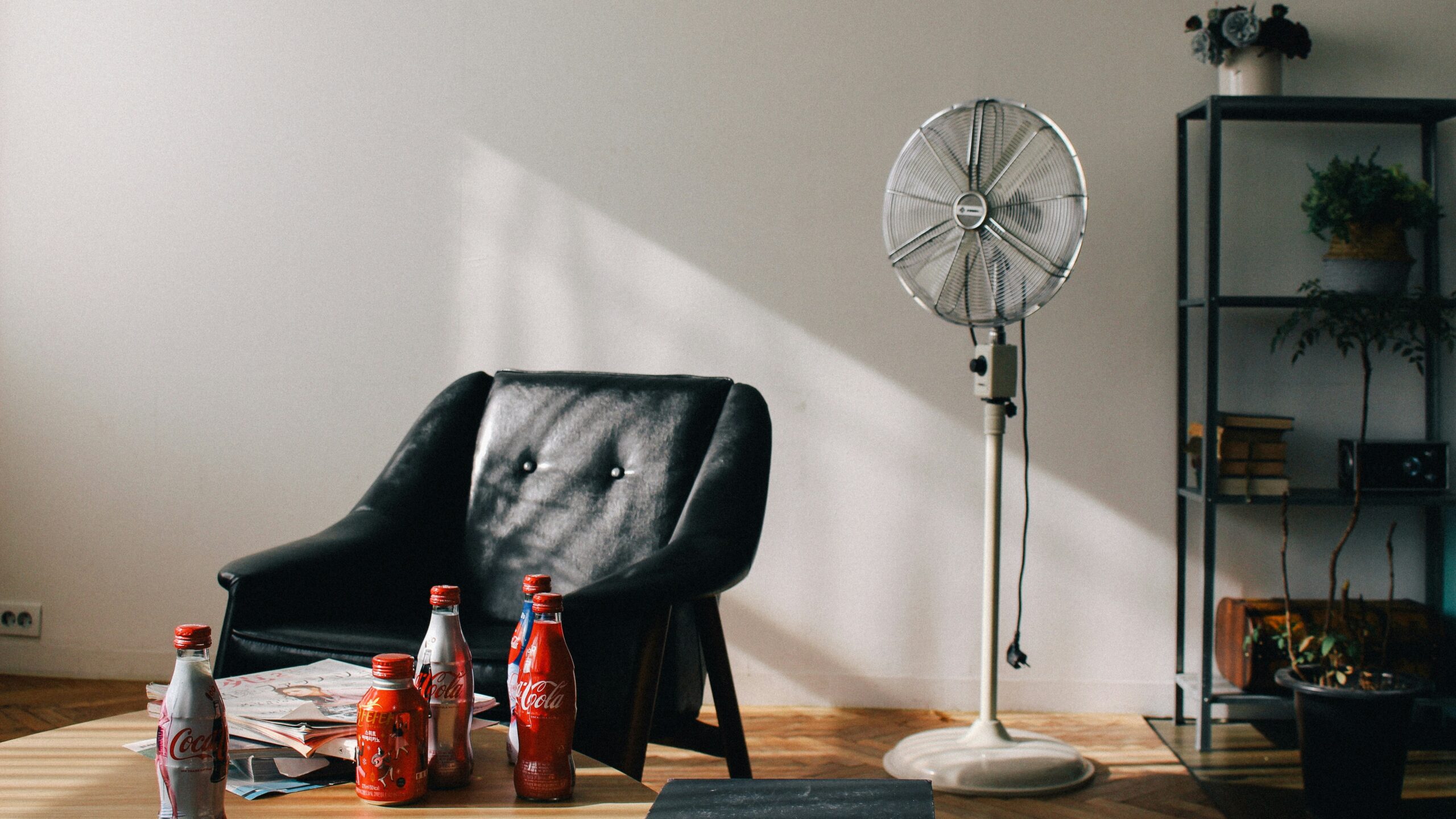 Coke soda bottles near vacant black armchair beside white pedestal fan inside white room