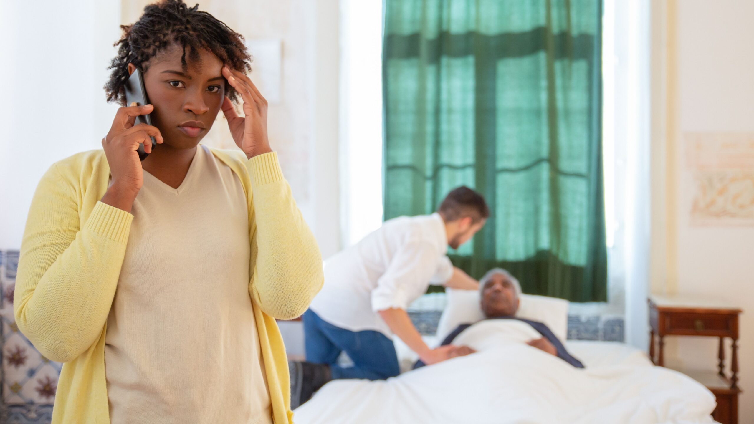 A woman on the phone looks worried as a caregiver assists an elderly person in bed.